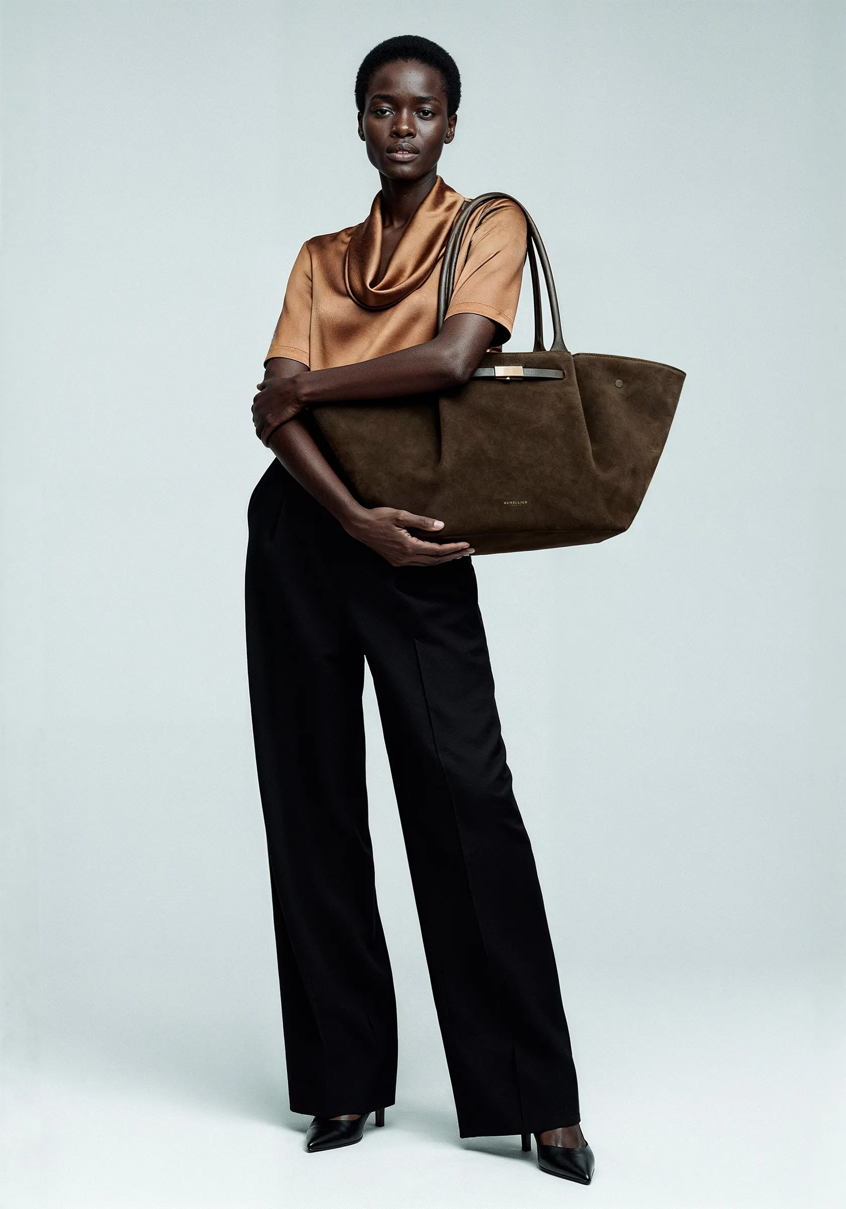 A model in a bronze blouse and black trousers carrying a suede tote